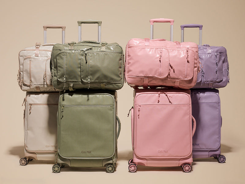 Set of colorful CALPAK Terra Collection backpacks and suitcases stacked on a beige background