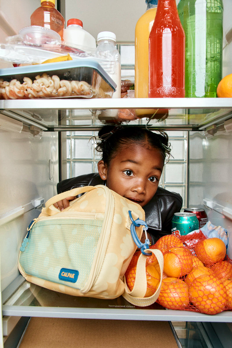 Child peeking into an open refrigerator filled with food items and CALPAK Kids Insulated Lunch Bag