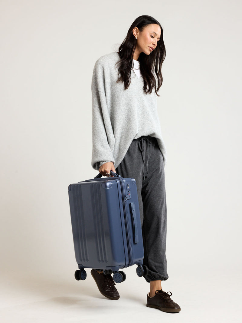 Model holding CALPAK Ambeur Carry On Luggage in dark blue metallic Indigo