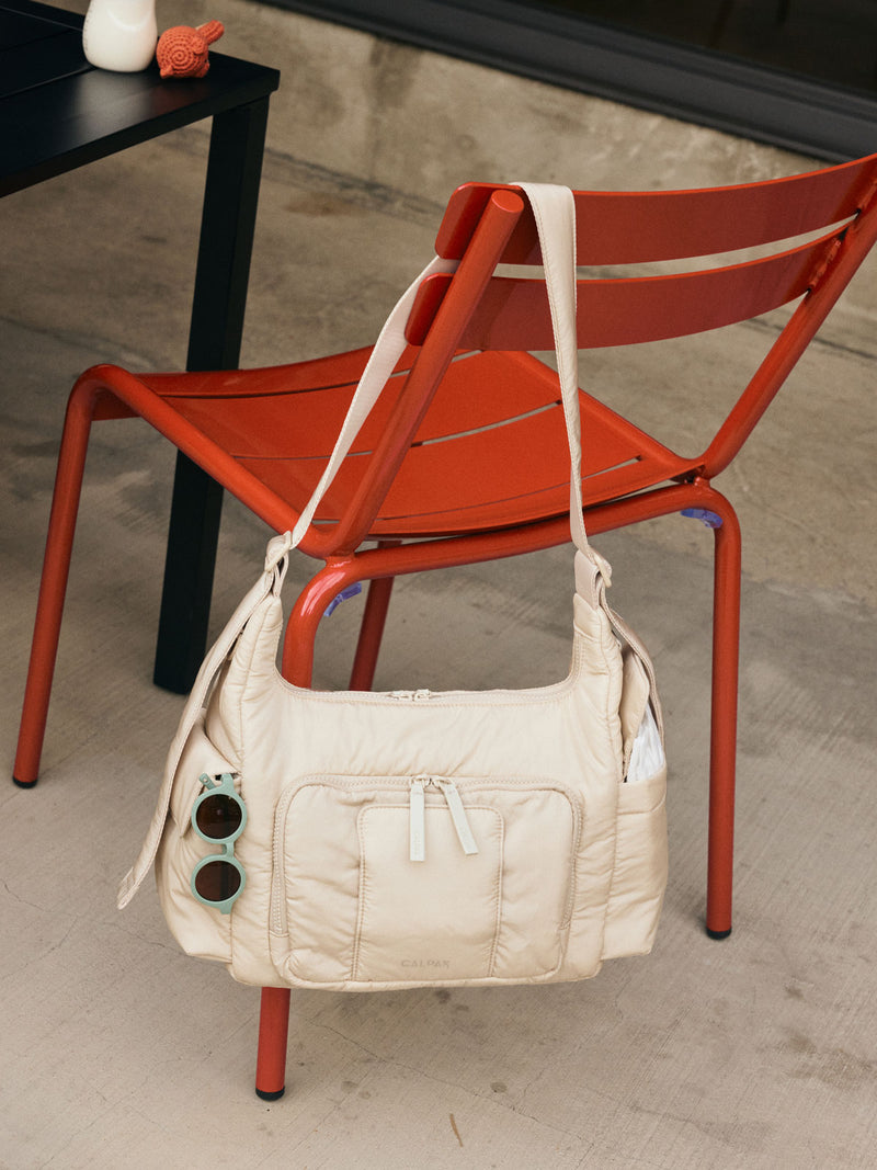 CALPAK Crossbody Diaper Bag in off-white Oatmeal hung on a red chair at a cafe