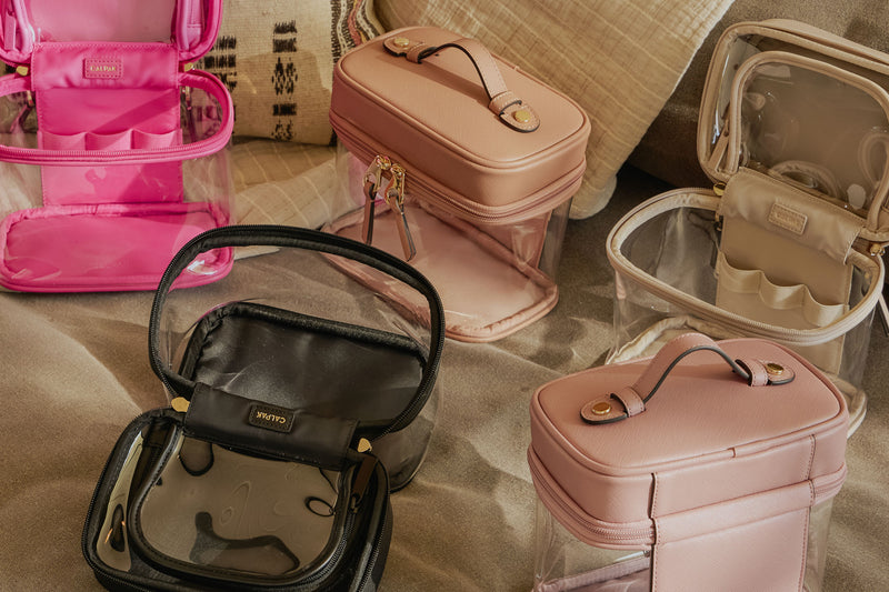 CALPAK Clear Train Cases in bright pink, pink, beige and black on the sand 