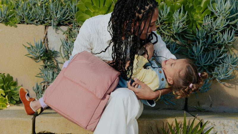 Woman holding a child with a pink CALPAK Diaper Bag outdoors