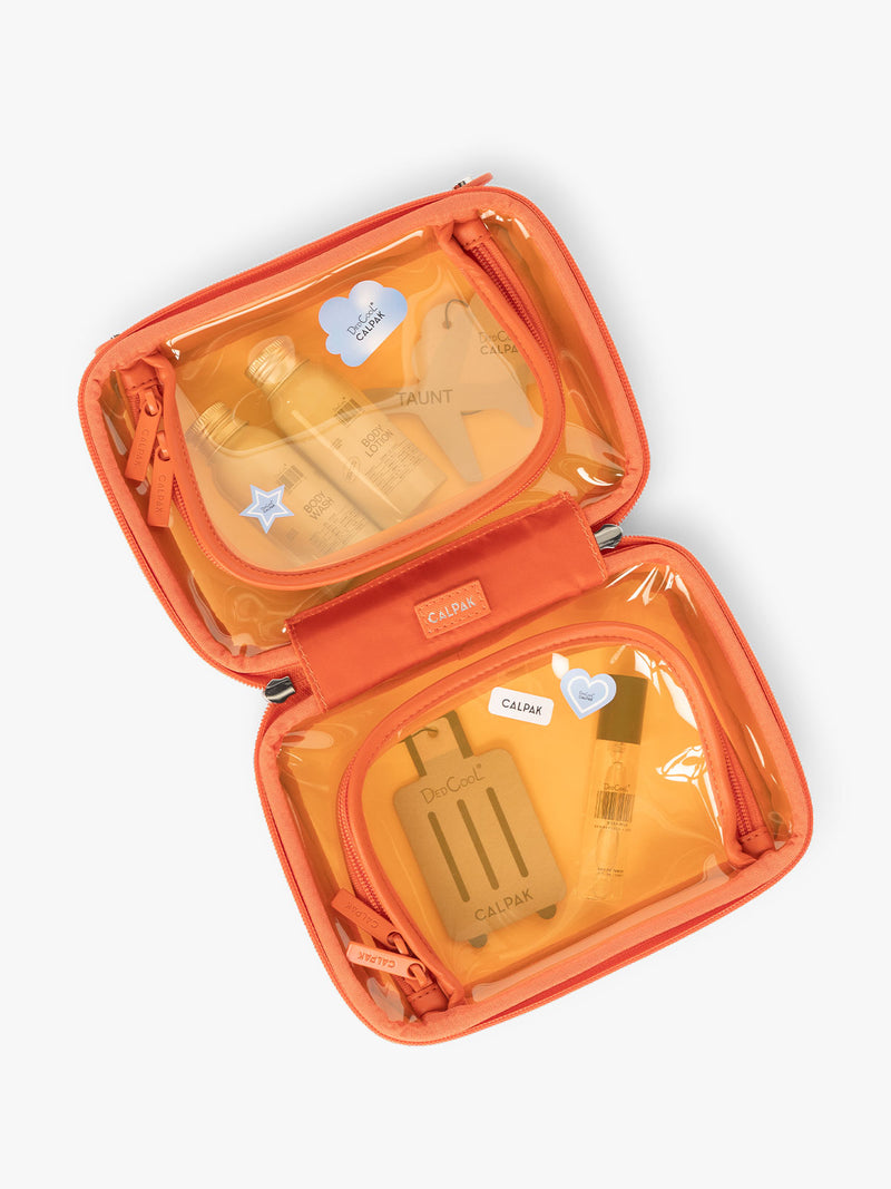 Opened view of CALPAK x Dedcool Collaboration of the Small Clear Cosmetic Case in orange Papaya with toiletry items and perfumes  on a airport chair