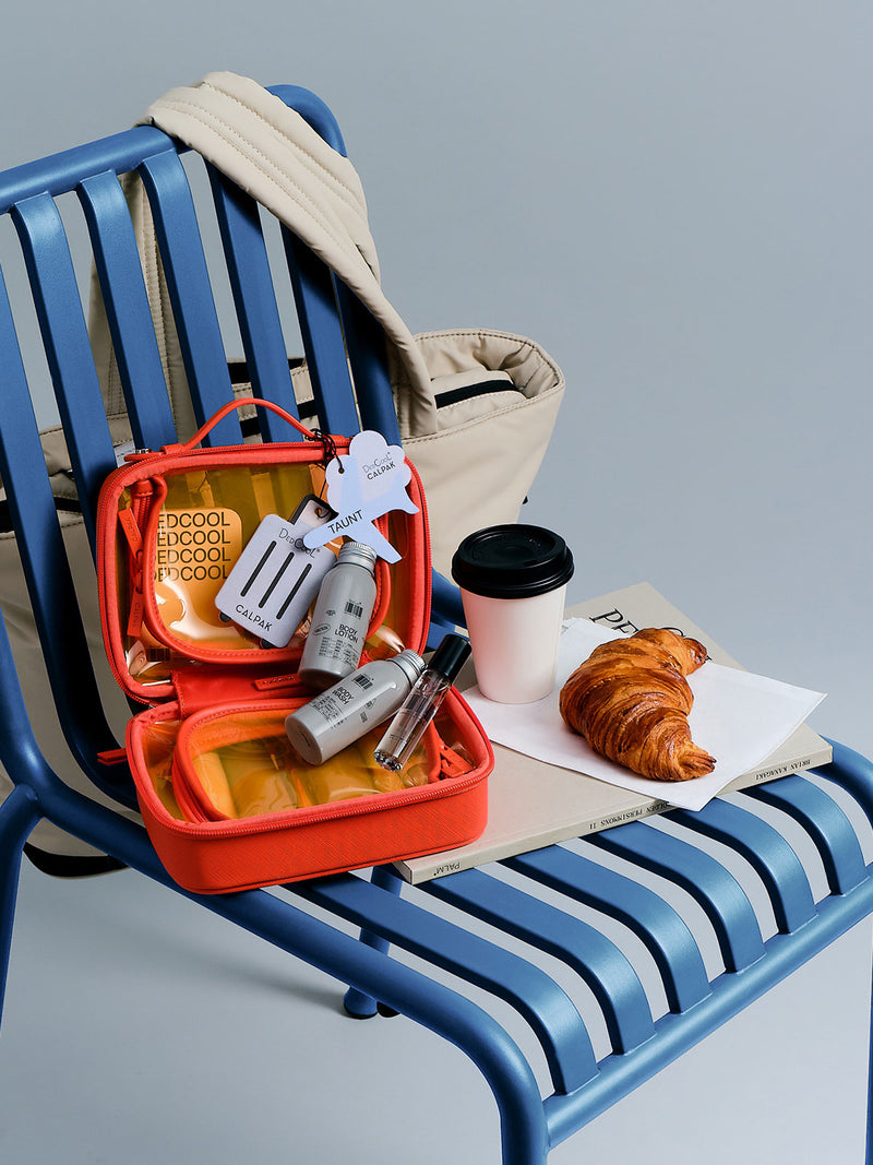 CALPAK x Dedcool Collaboration of the Small Clear Cosmetic Case in orange Papaya with toiletry items and perfumes  on a airport chair