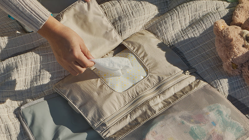 Person pulling a baby wipe from a CALPAK Diaper Changing Pad Organizer with a teddy bear and blanket in the background