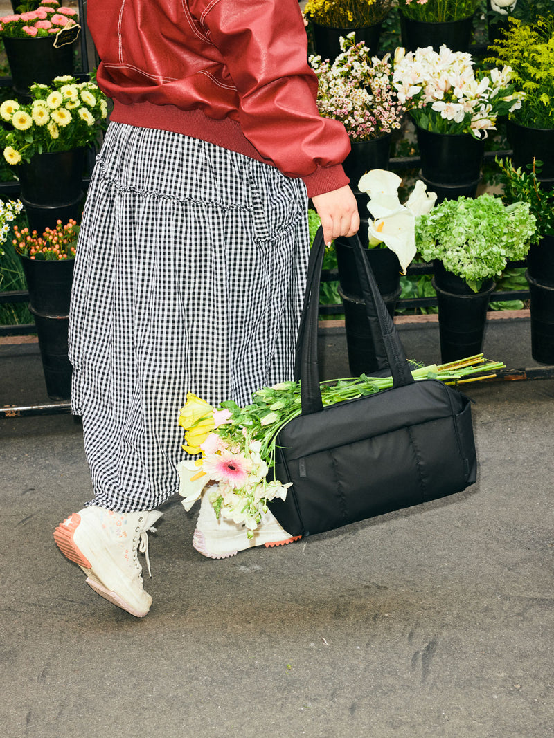 CALPAK Mini Duffel in Black with puffy exterior, interior and exterior pockets, and padded shoulder straps being held by a model with flowers
