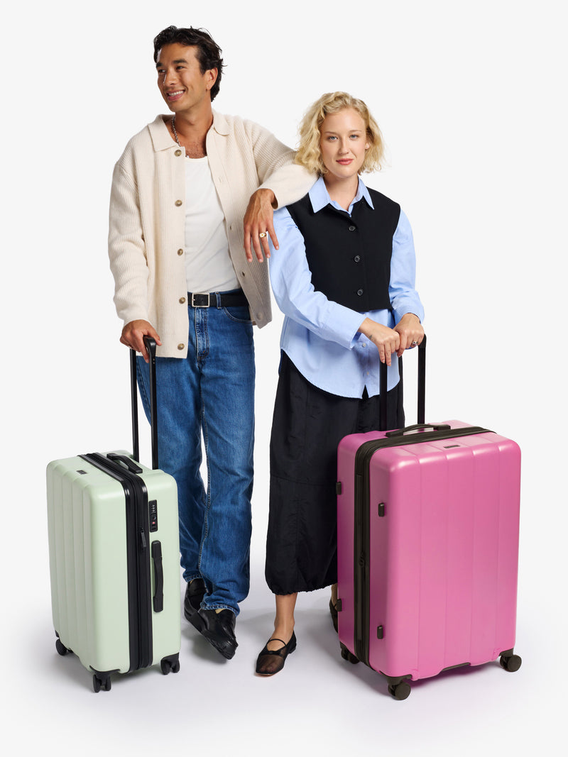 Man posing with a green small carry-on suitcase next to a woman posing with a pink large suitcase, showcasing stylish luggage options