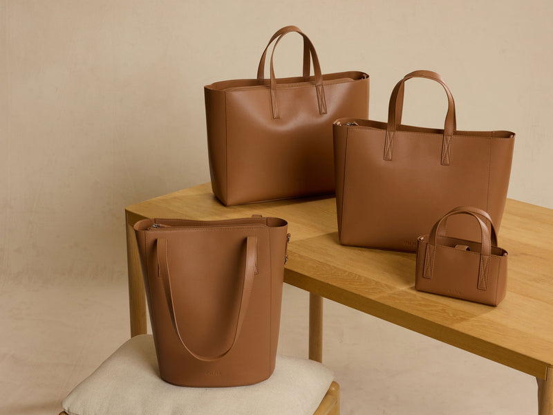 Four brown leather CALPAK Haven Collection handbags on a wooden table against a beige background