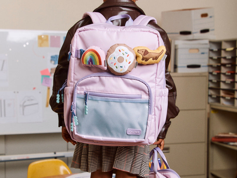 Person wearing a pink CALPAK backpack with decorative Pluffy Bag Charms in an indoor setting