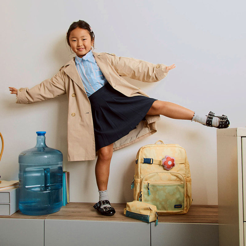 Child in a trench coat with a CALPAK Kids Collection backpack and water bottle on a shelf