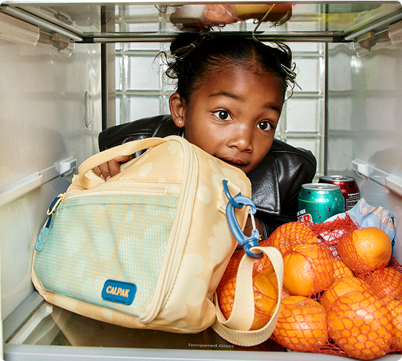 Child peeking into an open refrigerator with a Calpak bag and oranges.
