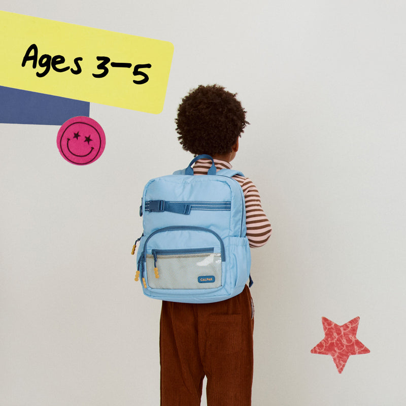 Child wearing a Small CALPAK blue backpack with colorful text and shapes on a light gray background