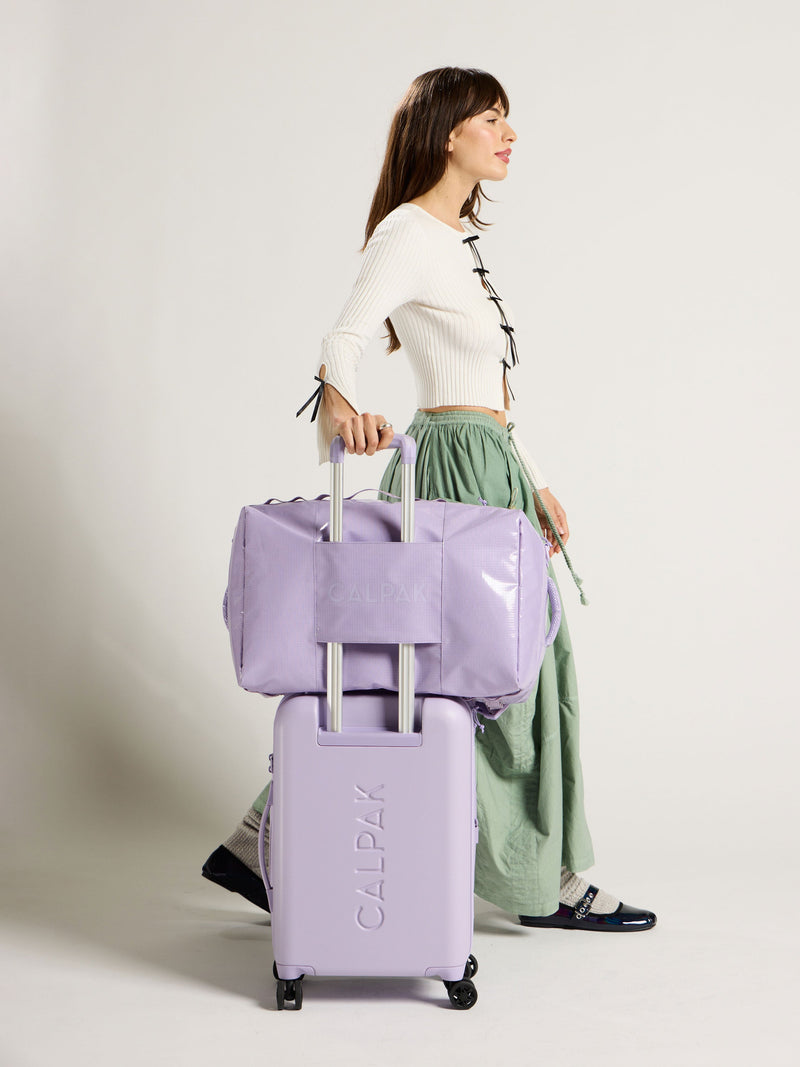 Model rolling CALPAK Terra 46L Carry-On Luggage with CALPAK Terra Large 50L Duffel Backpack on trolley passthrough in light purple; DTL2301-AMETHYST