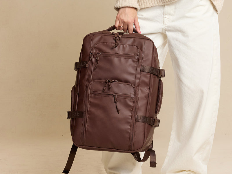 Brown CALPAK Terra 26L Laptop Duffel travel backpack held by a person against a beige background