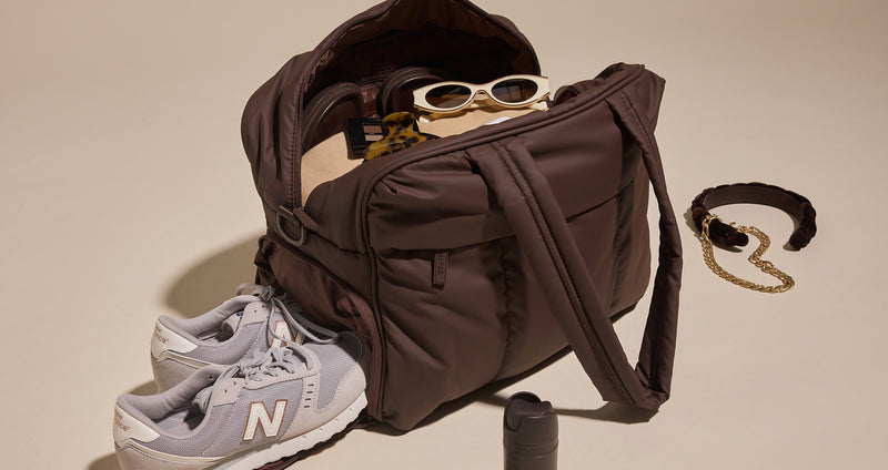 Brown CALPAK Luka Duffel with sunglasses, gray sneakers, and a necklace on a beige background