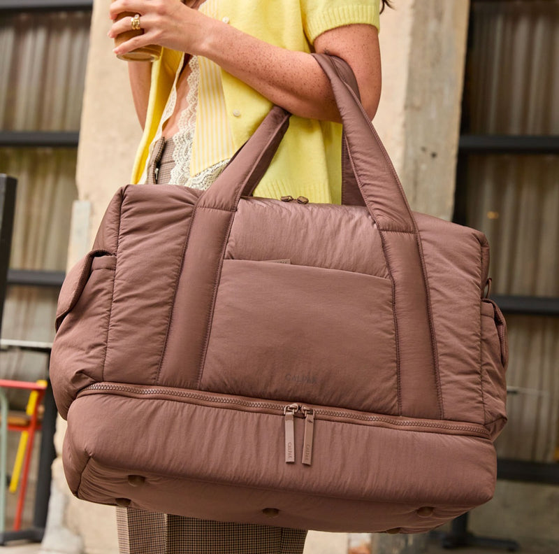 Brown quilted CALPAK Weekender Duffel handbag held by a person wearing a yellow top.