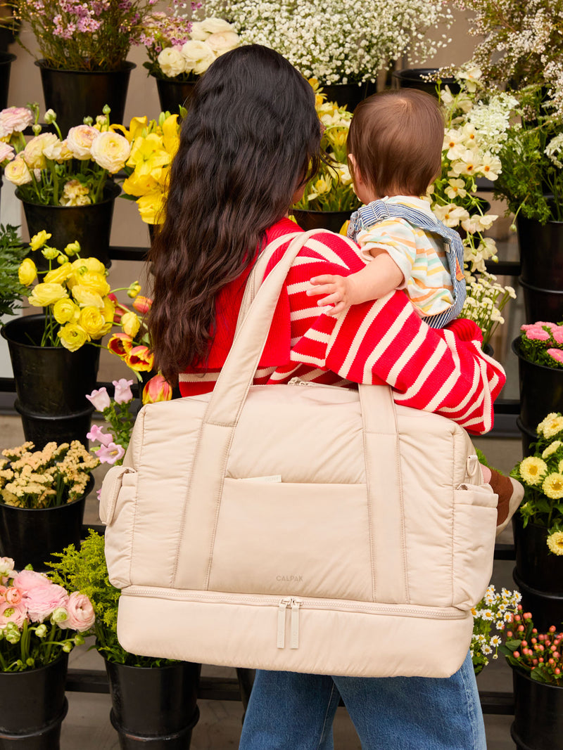 Model carrying baby and wearing the CALPAK Weekender Duffel in white Oatmeal; WDB2501-OATMEAL