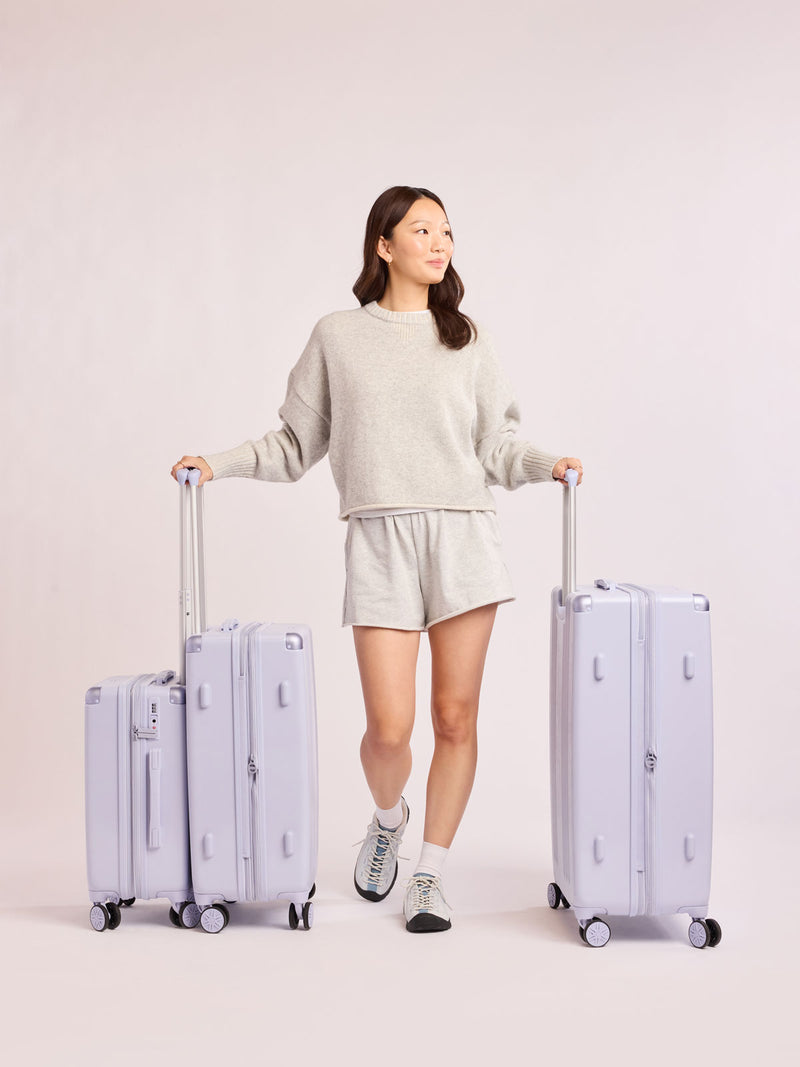 Woman holding the CALPAK Ambeur three piece luggage set in carry on, medium, and large sizes in light purple lavender with handles and spinner wheels on a white background