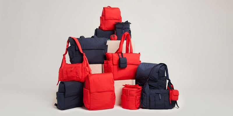 Stack of red and navy CALPAK Baby Collection bags and accessories on a light gray background