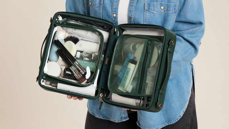 Person holding a green Emerald CALPAK Clear Cosmetics Case with toiletries on a beige background
