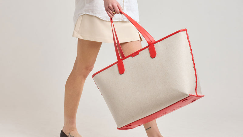 Person holding a cream and blue CALPAK Colette Canvas Tote in beige and red Tomato handbag with a red strap against a plain background
