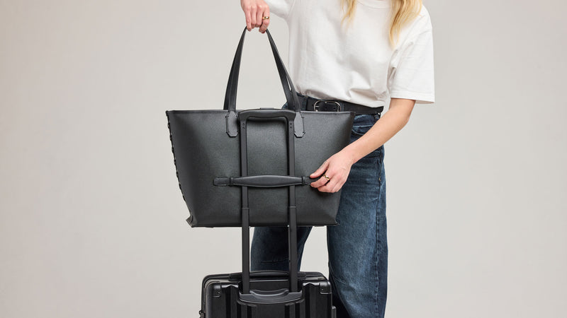 Person holding a CALPAK Canvas Colette Tote Bag in Black over a suitcase on a plain background