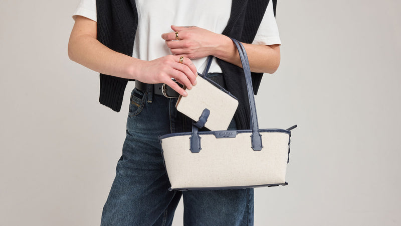 Person holding a CALPAK Colette Canvas Shoulder Tote in beige and Navy handbag with a navy strap against a plain background
