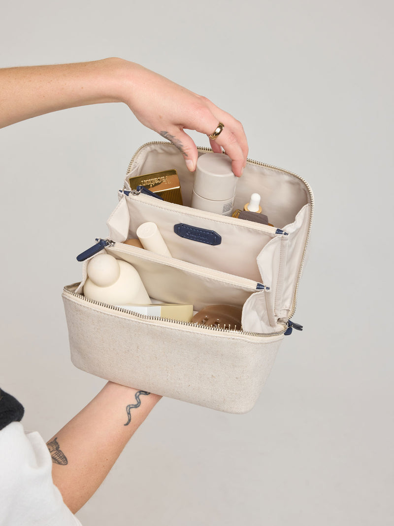 Woman putting items in CALPAK Colette Lay-Flat Canvas Toiletry Bag in beige canvas material and blue finishing