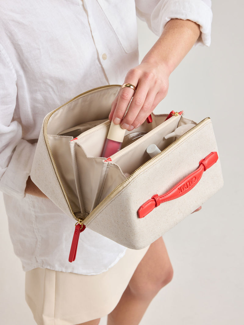 Model putting item into CALPAK Colette Lay-Flat Canvas Toiletry Bag in beige canvas material and red finishing