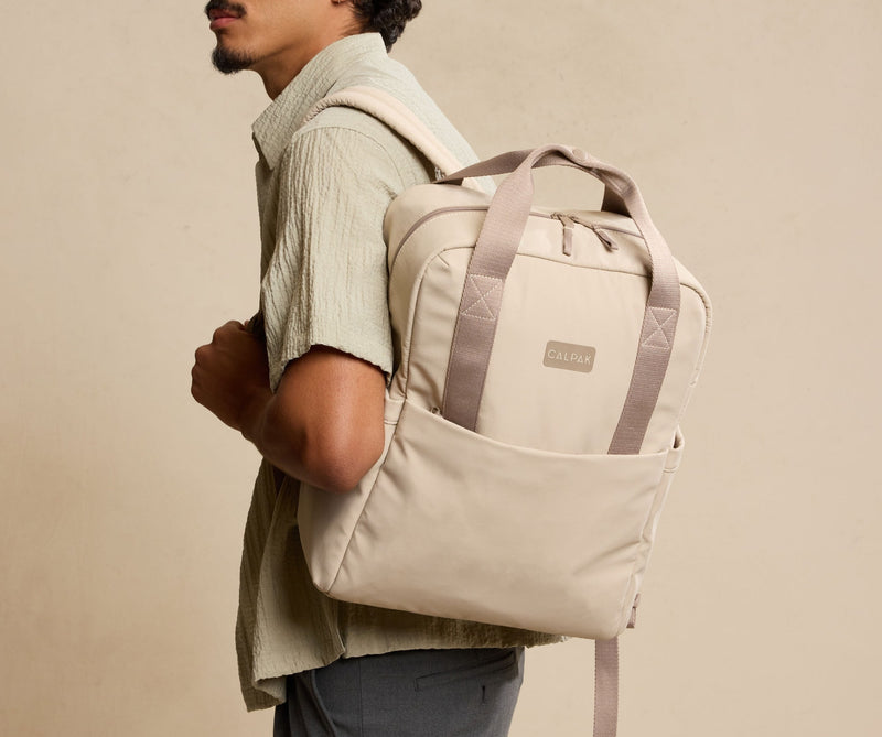 Man wearing a beige CALPAK Connect Laptop backpack with a visible CALPAK brand logo on a beige background