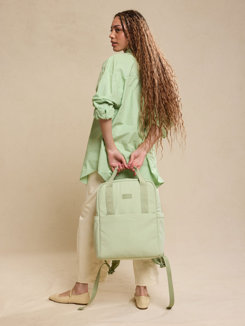 CALPAK Connect Laptop Backpack in light green Meadow being held by model front view on a beige background