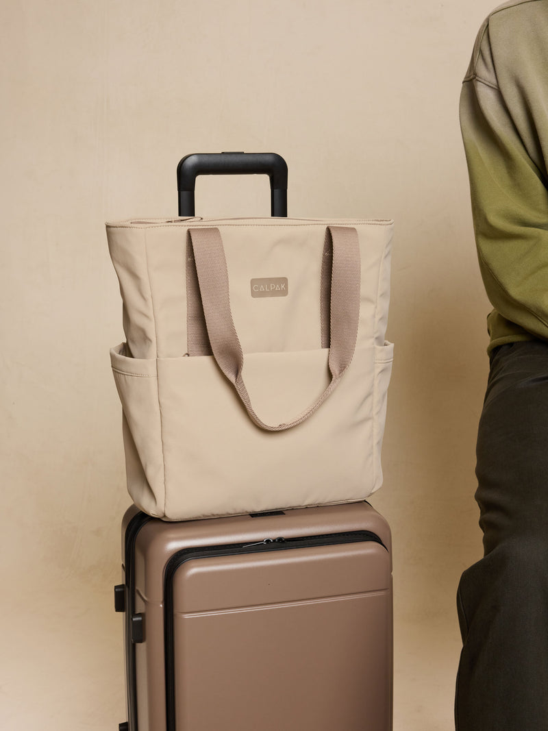 CALPAK Zippered Laptop Tote Bag in beige Greige on a suitcase against a beige background