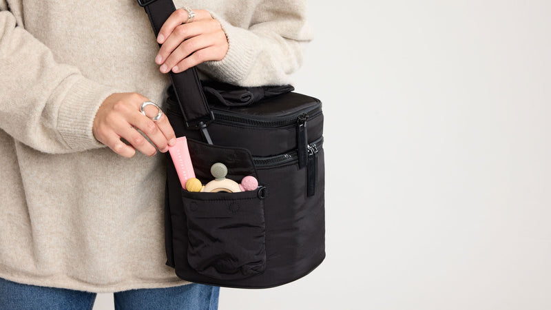Person opening pocked of a black CALPAK Insulated Baby Bag with items inside on a light gray background