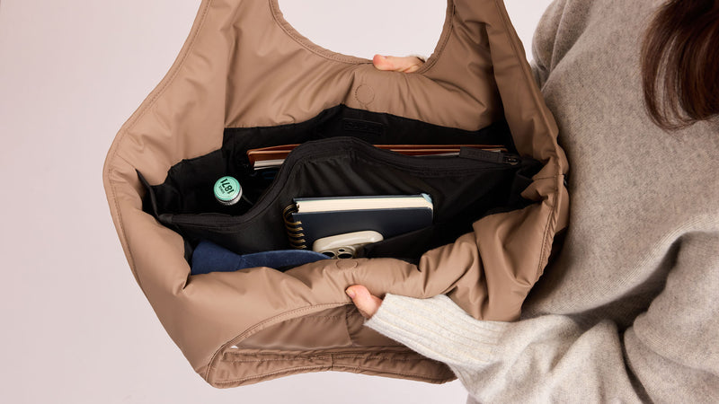 Person holding the Luka Everything Tote in brown Chocolate with items inside on a light pink background