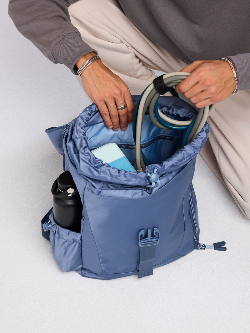 Person adding gym essentials to the CALPAK Fit Gym Backpack in blue Indigo