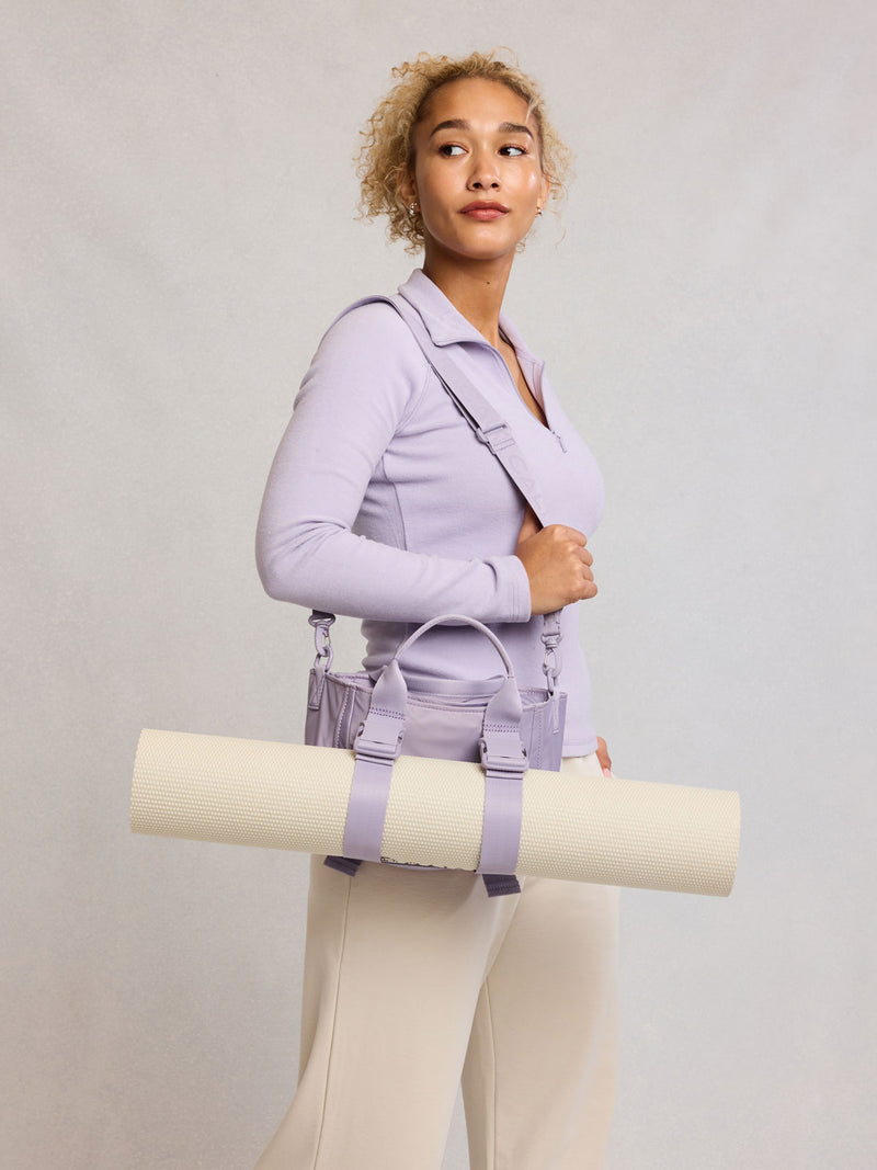 Model wearing CALPAK Fit Gym Crossbody Bag in light purple Iris with Yoga mat attached to front