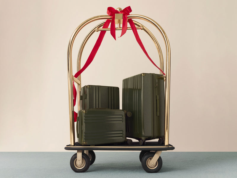 Gold luggage cart with CALPAK Ambeur green suitcases and a red ribbon on a beige background