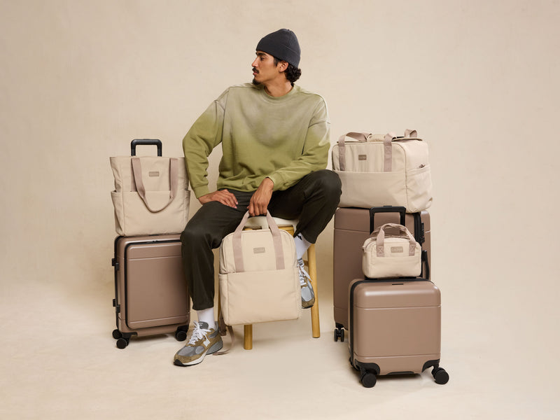 Man sitting on a stool surrounded by various CALPAK Connect Collection bags and Hue Collection suitcases on a beige background