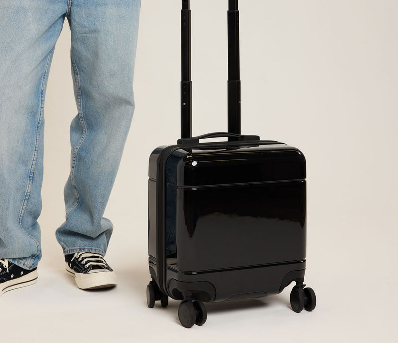 Black CALPAK mini carry-on suitcase with wheels and handle next to a person wearing jeans and sneakers on a light background