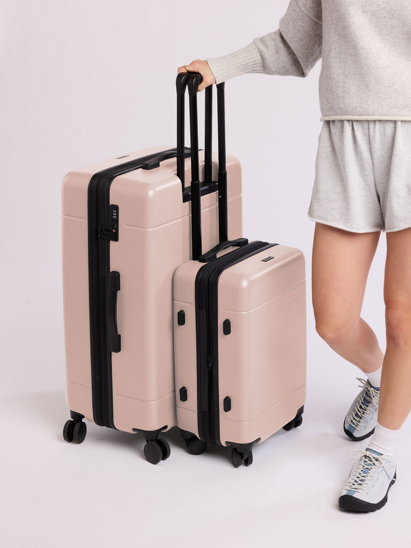 Person holding a set of rose quartz CALPAK luggage part of Hue Starter Bundle with black handles and wheels on a light gray background