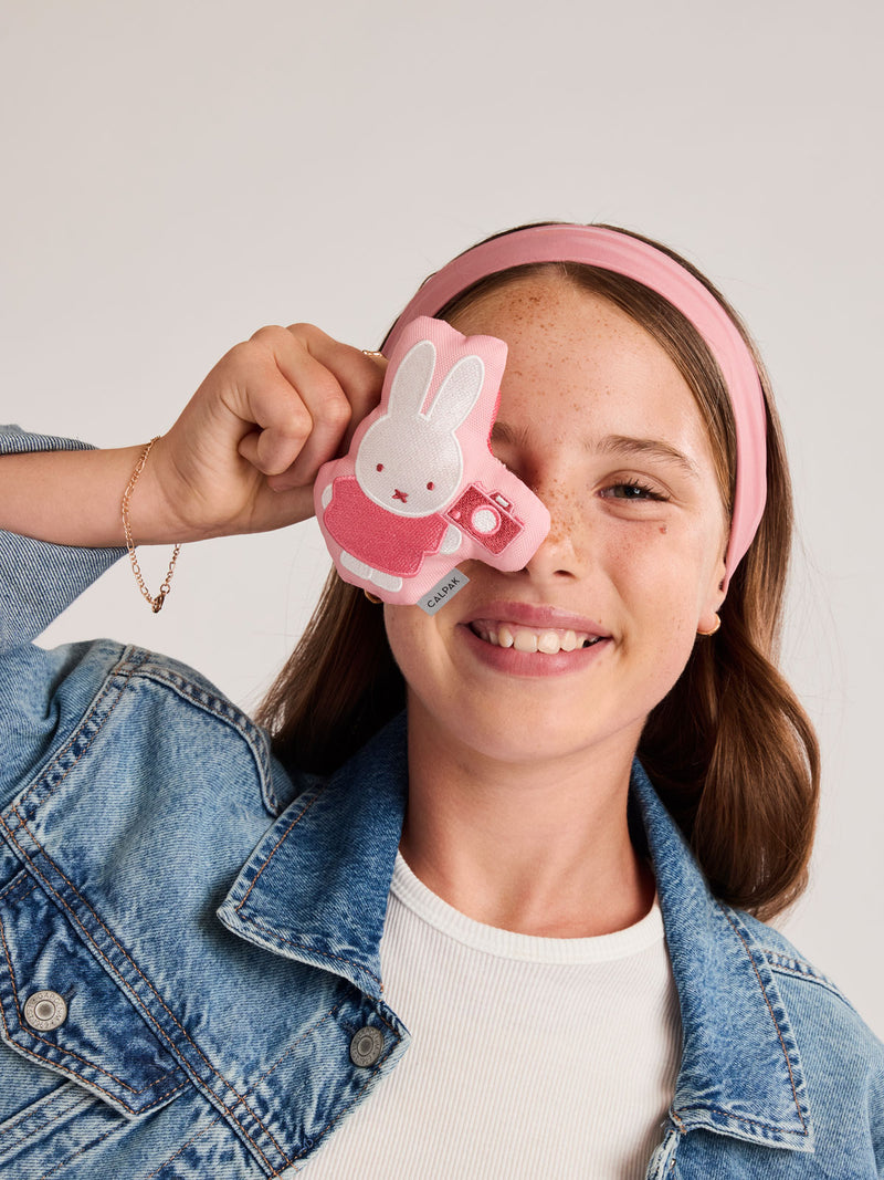 Girl posing with the Miffy x CALPAK Pluffy Bag Charm keychain with a pink border and Miffy holding a camera