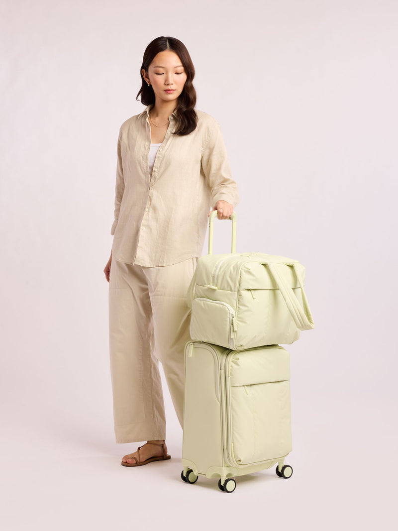 Model with CALPAK Luka Soft-Sided Carry On Luggage in light green Celadon on a white background with handle and spinner wheels