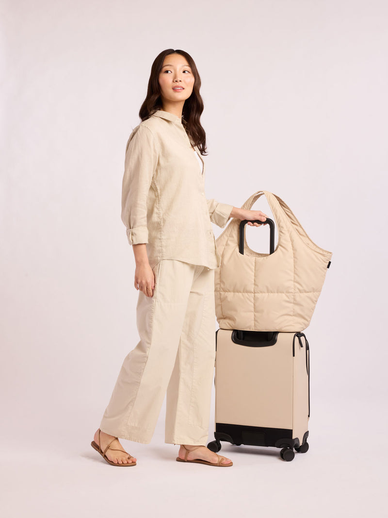 Model with CALPAK Luka Everything Tote in off-white beige Oatmeal on top of a suitcase with trolley luggage sleeve