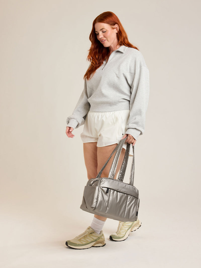 Model holding CALPAK Luka Mini Duffel in metallic grey on a white background