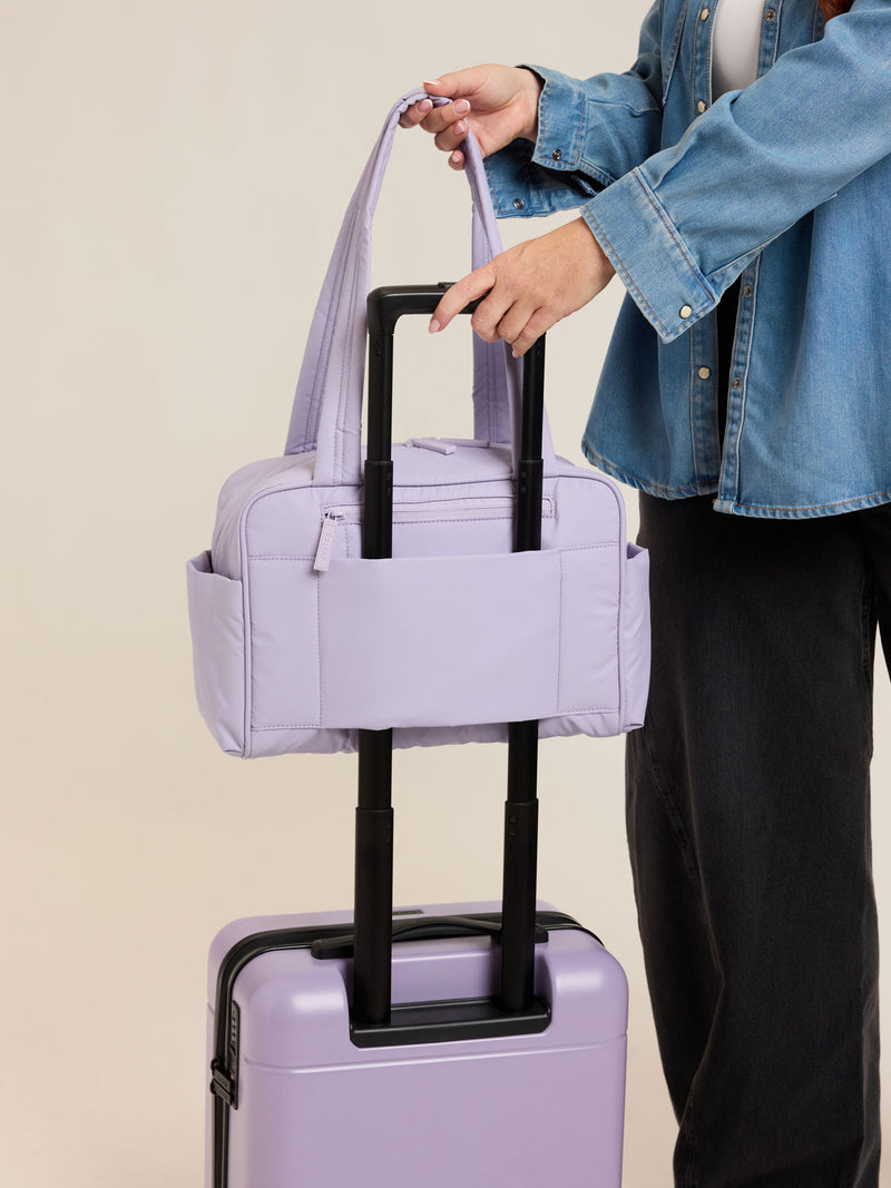Model using trolley sleeve of CALPAK Luka Mini Duffel in light purple Orchid on a white background
