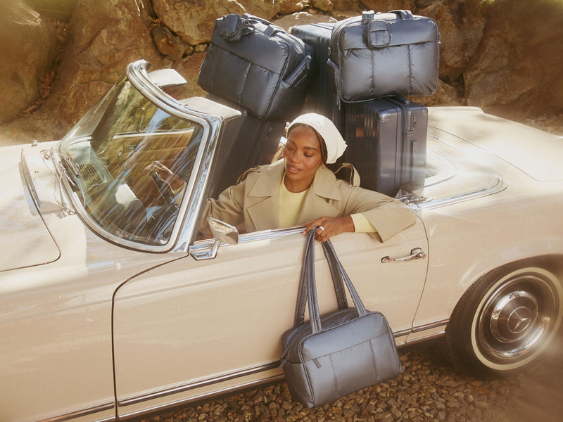 Woman with metallic indigo Ambeur luggage and Luka bags in a vintage convertible car on a rocky road