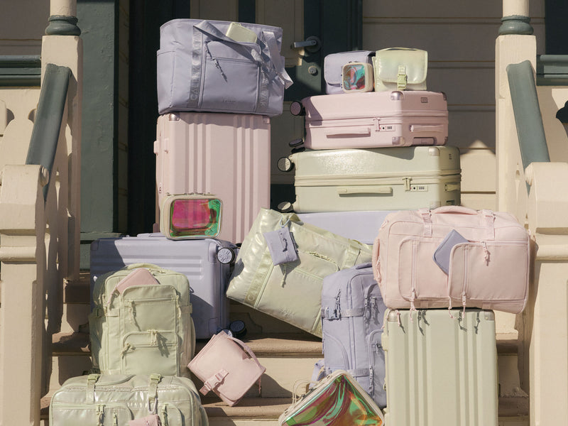 Stack of pastel-colored CALPAK Ambeur suitcases and Terra bags on a wooden balcony.