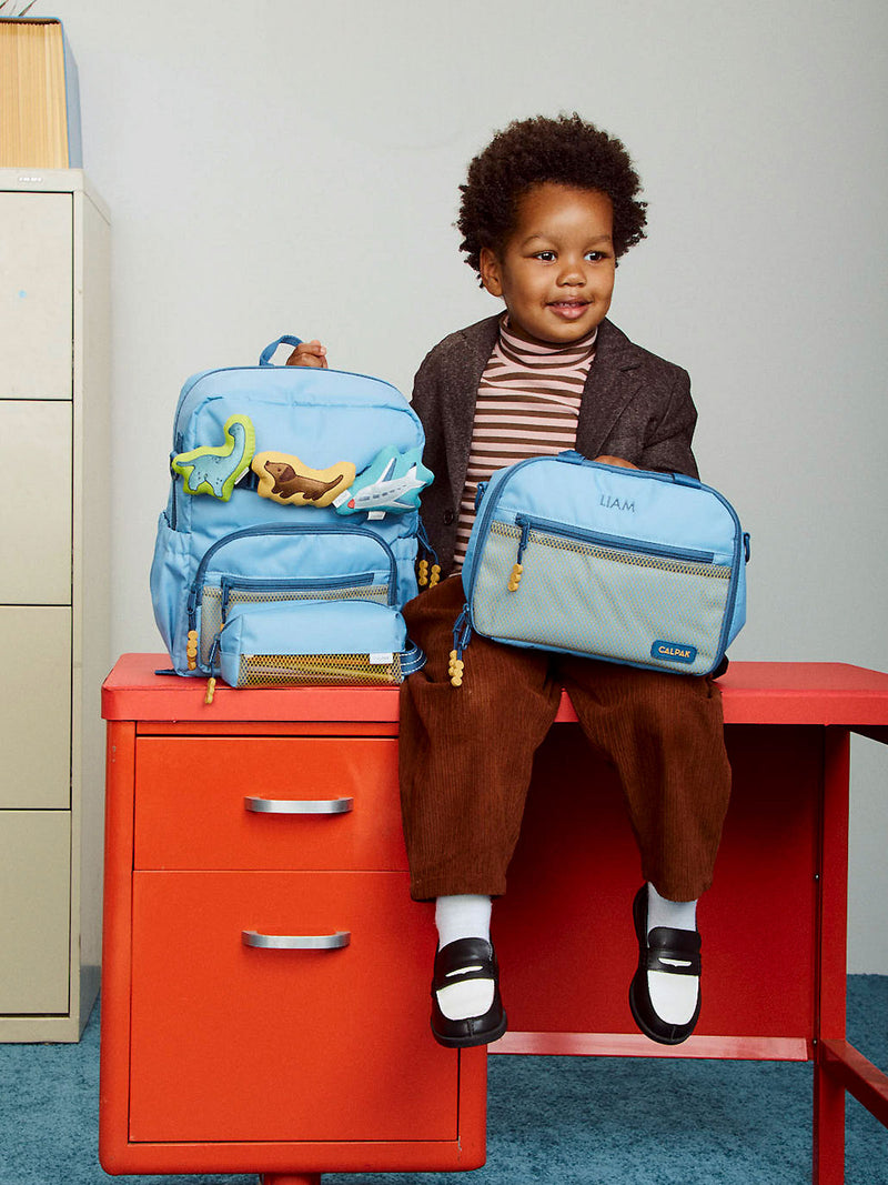 CALPAK Kids Small Backpack in Blue with a blue Dinosaur, brown Dog, and white Airplane Pluffy Bag Charms on a school desk with child holding a lunch bag