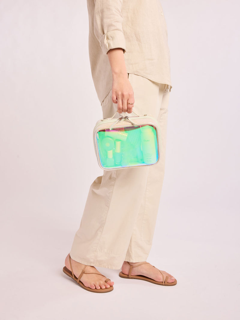 Person wearing beige holding a CALPAK Small Clear Cosmetic Case in Moonbeam with white border and transparent holographic iridescent makeup bag with pink on a white background