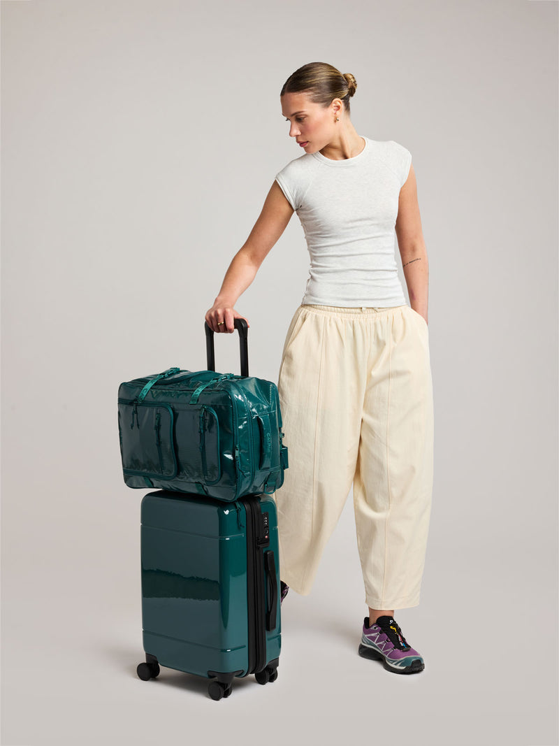 Model with the CALPAK Terra 26L Laptop Backpack in dark blue green Kale with multiple pockets on top of a suitcase